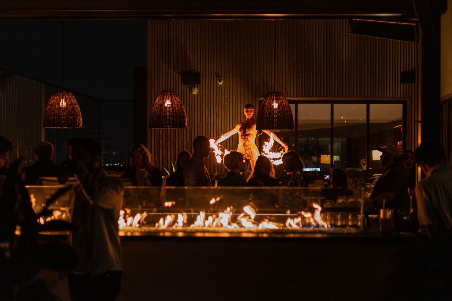 People gather around a long fire pit at night with warm ambient lighting from pendant lamps in a cozy restaurant setting.