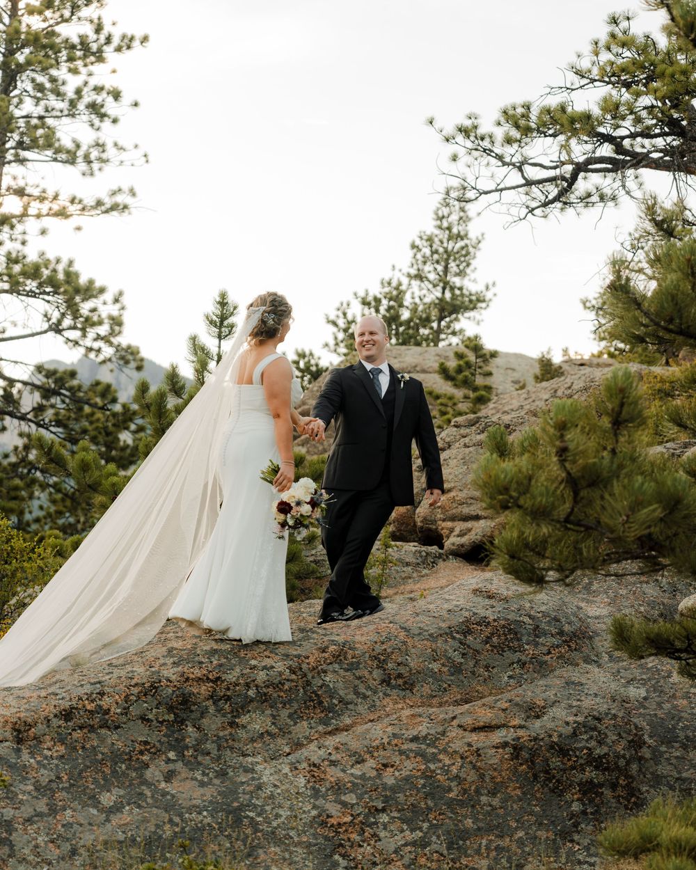Bride and groom holding hands on rocky mountain overlook surrounded by pine trees during outdoor wedding ceremony.