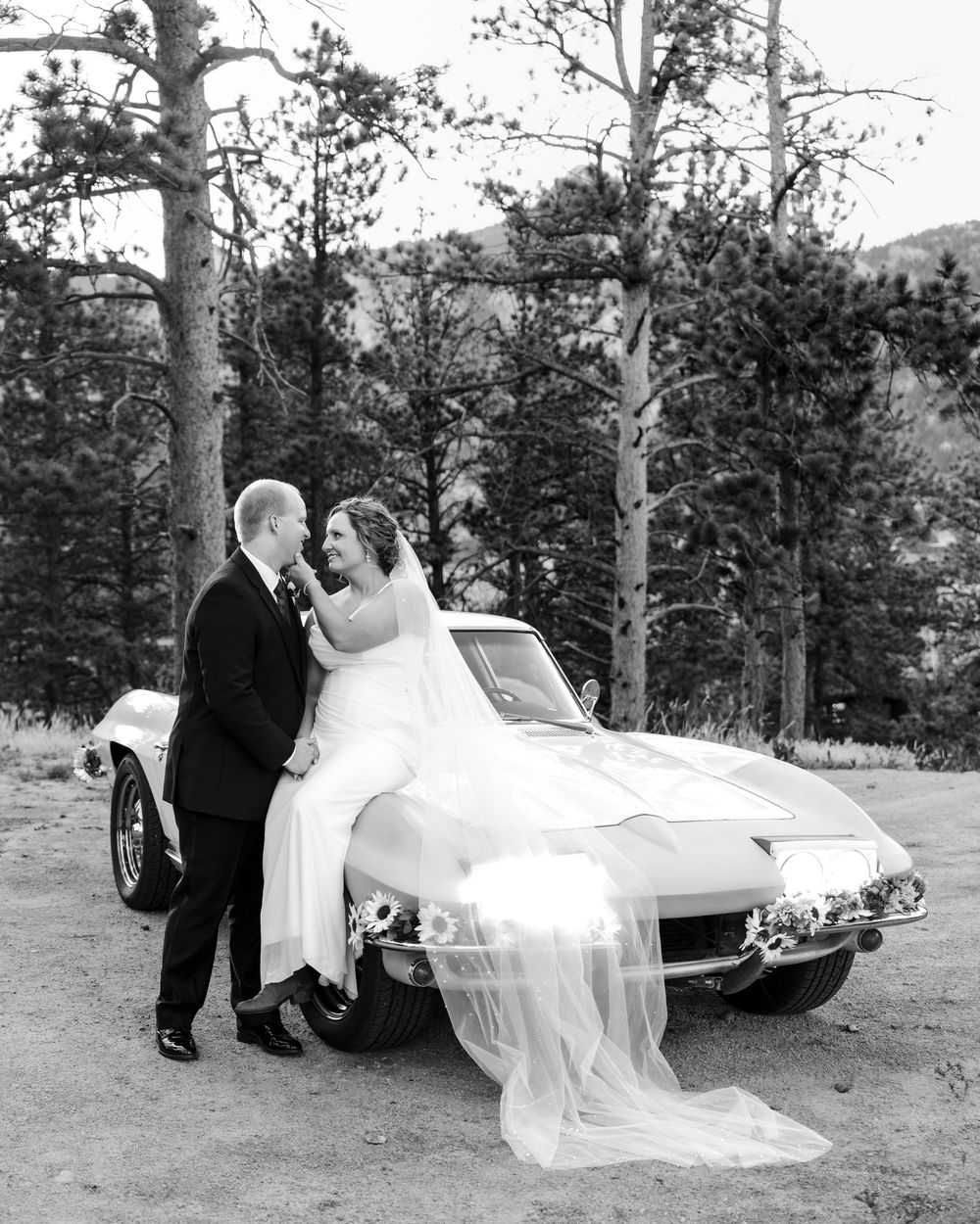 Black and white photo of bride sitting on classic Corvette hood with groom standing beside her, surrounded by pine trees.