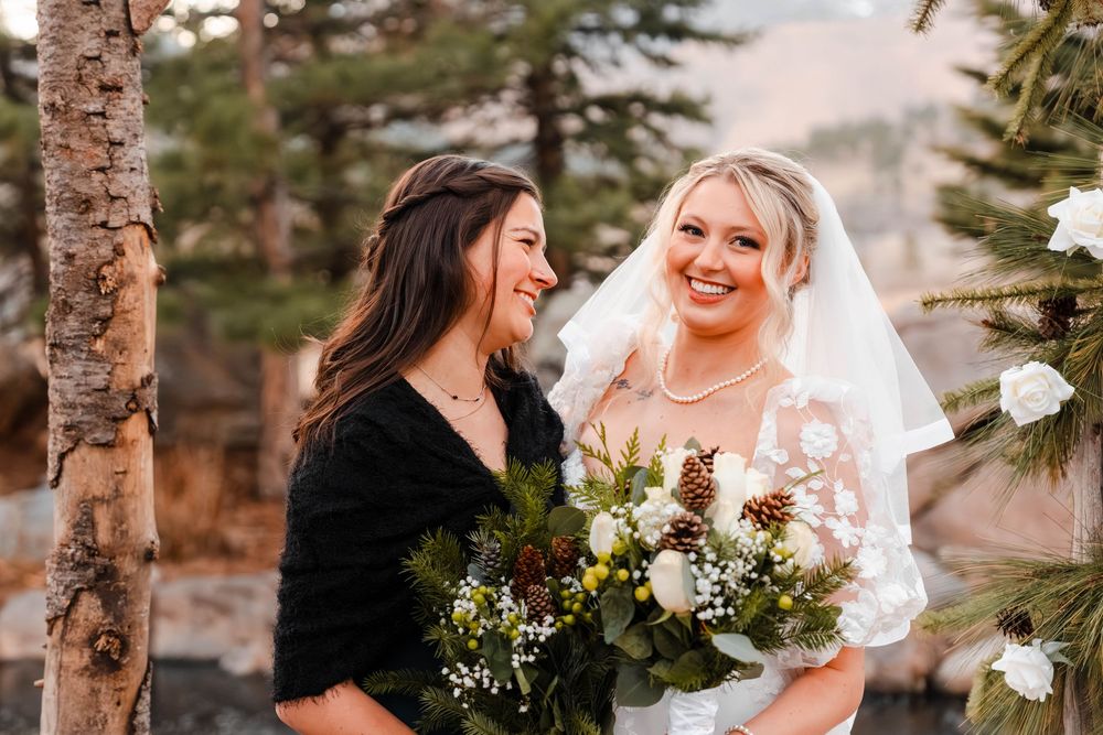 Bride in floral veil smiles with bridesmaid holding lush winter greenery bouquet outdoors.