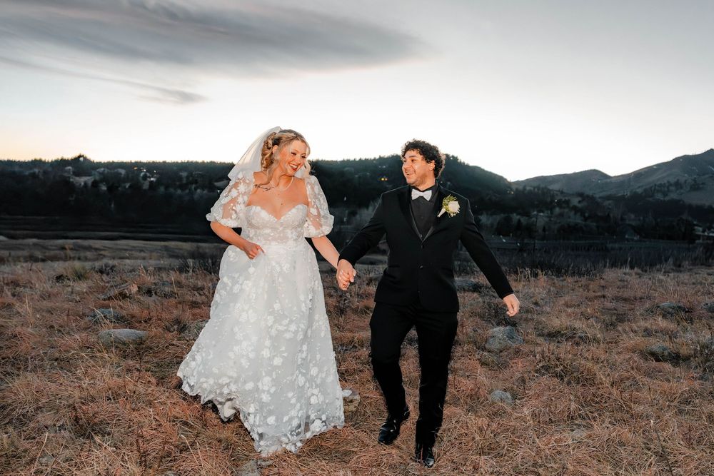 Bride and groom hold hands walking through golden mountain meadow at sunset after wedding.