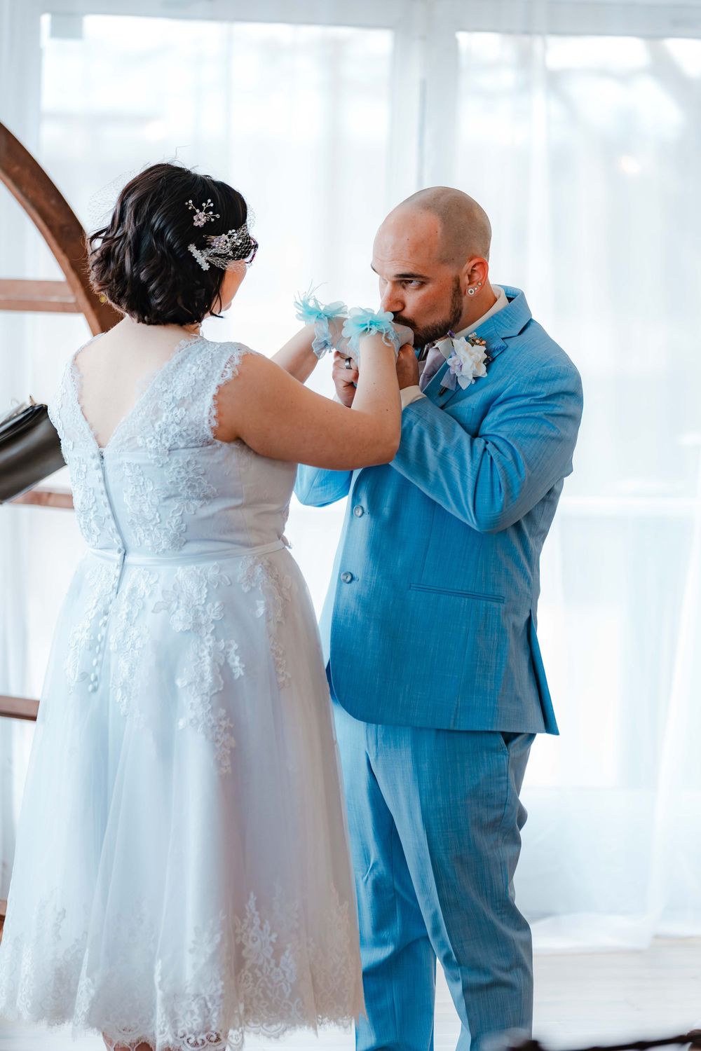 Groom in blue suit kisses bride's hand during wedding ceremony, bride wearing white lace dress.