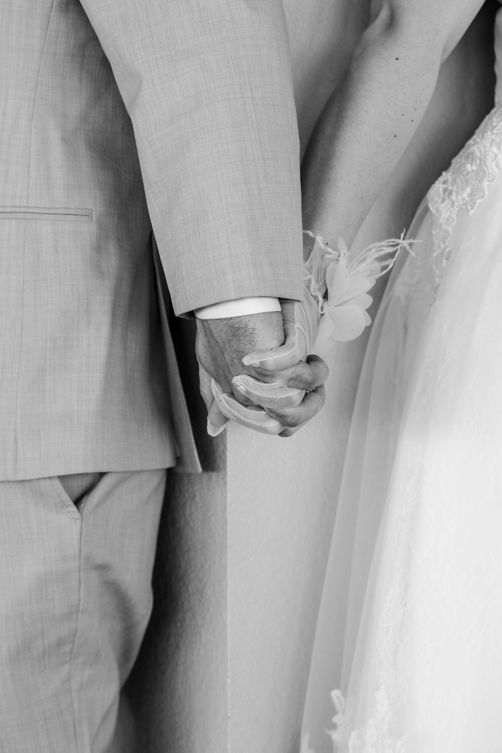 Black and white photo of a bride and groom holding hands, showcasing wedding attire and a delicate floral wrist corsage.