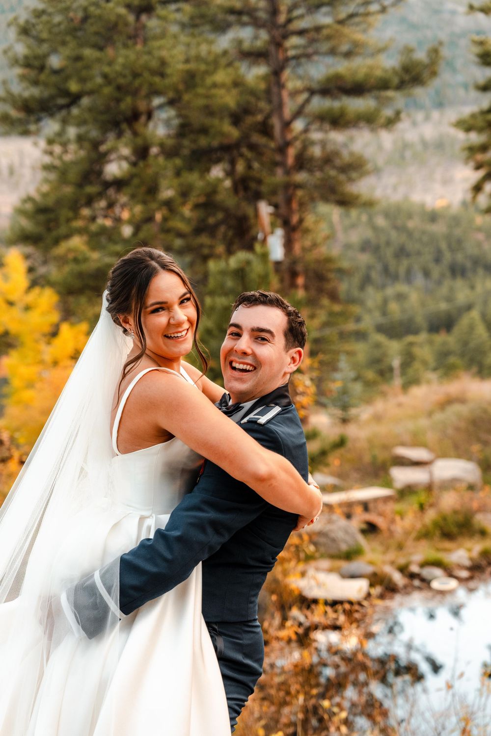 Laughing bride and groom embracing outdoors with colorful autumn mountain landscape behind them.