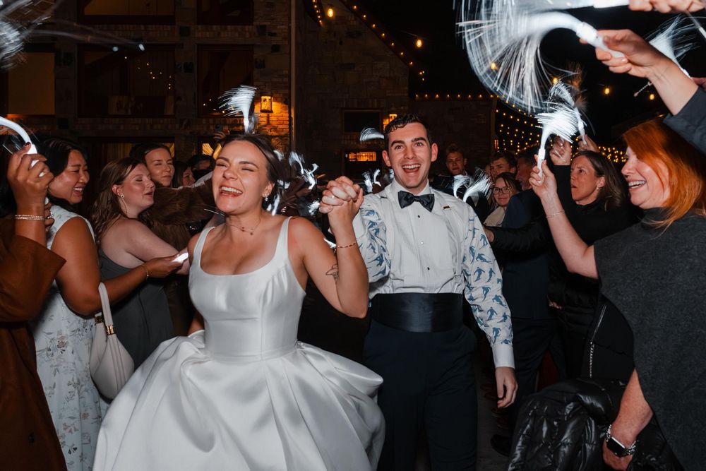 Bride and groom celebrating joyfully with guests waving white feather wands at wedding reception.