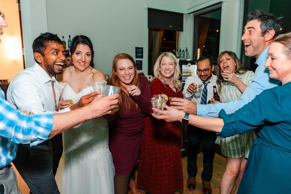 Bride groom and guests laugh and raise glasses in a celebratory toast at wedding reception.