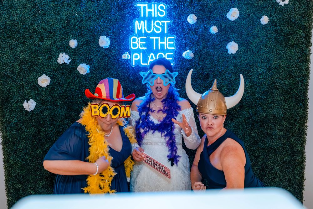 Three guests pose in fun costumes and props at a wedding photo booth with neon sign backdrop.