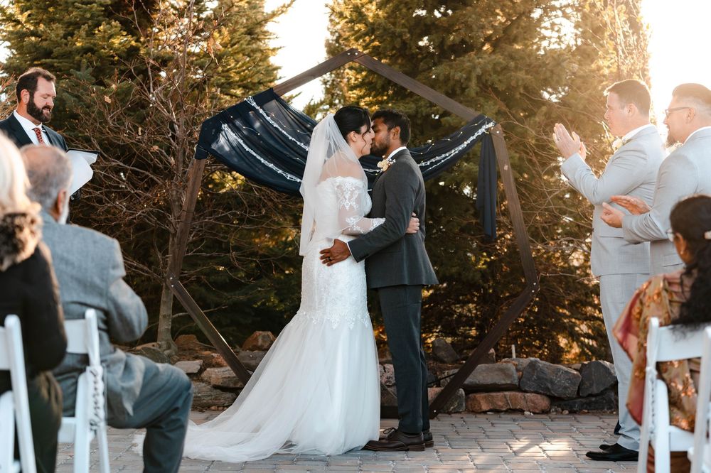 Bride and groom share first kiss at outdoor wedding ceremony with pine trees and golden sunset.