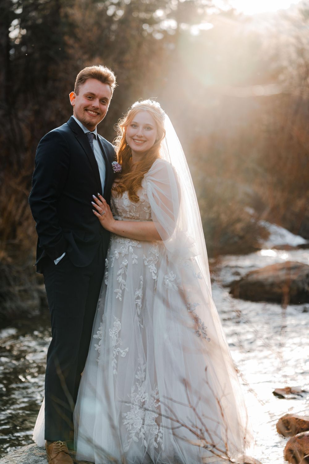 Bride and groom embracing by a sunlit river, bride in lace gown with veil, groom in navy suit.