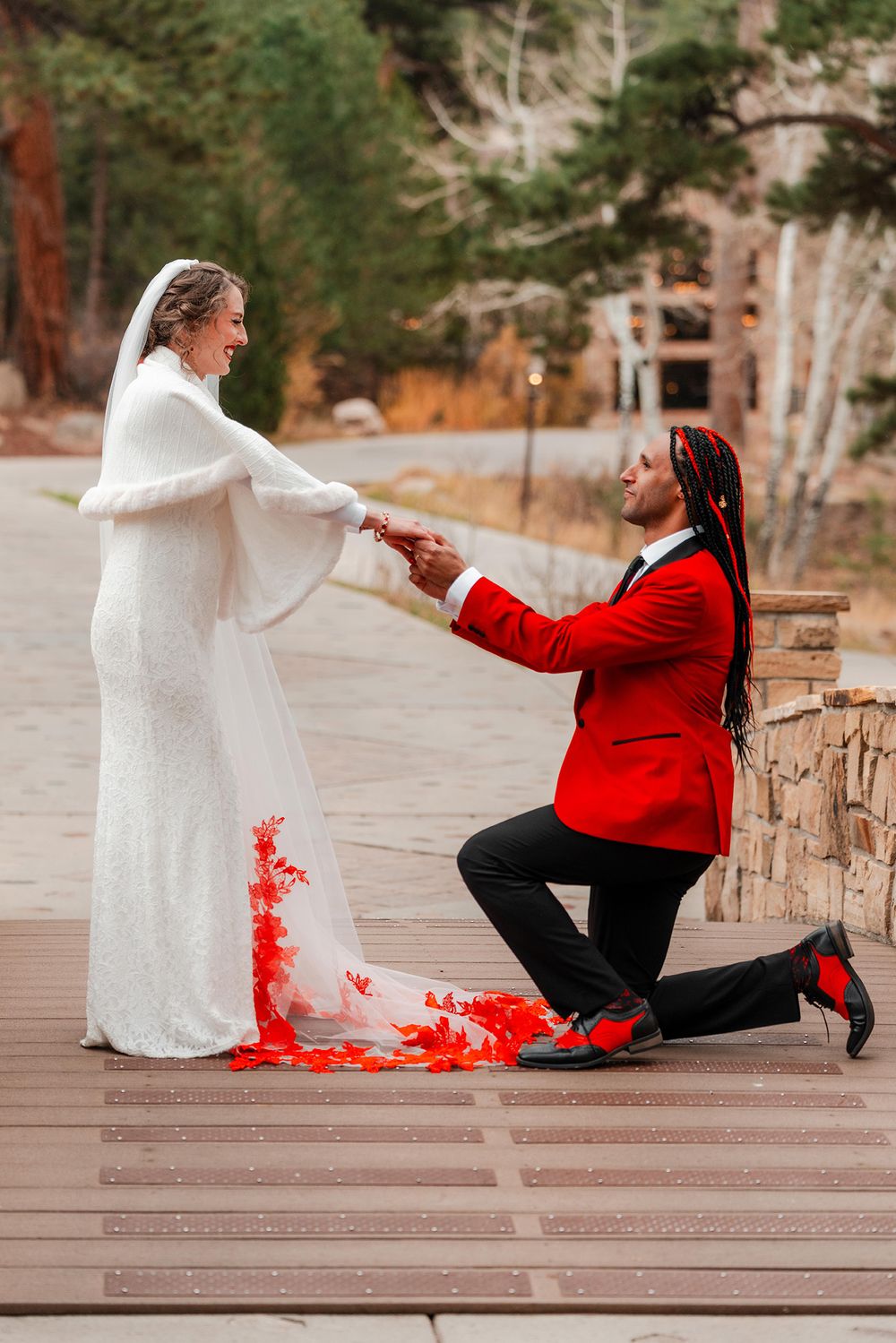 Groom in red tuxedo kneels before bride in white gown with red floral accents on wooden deck outdoors.