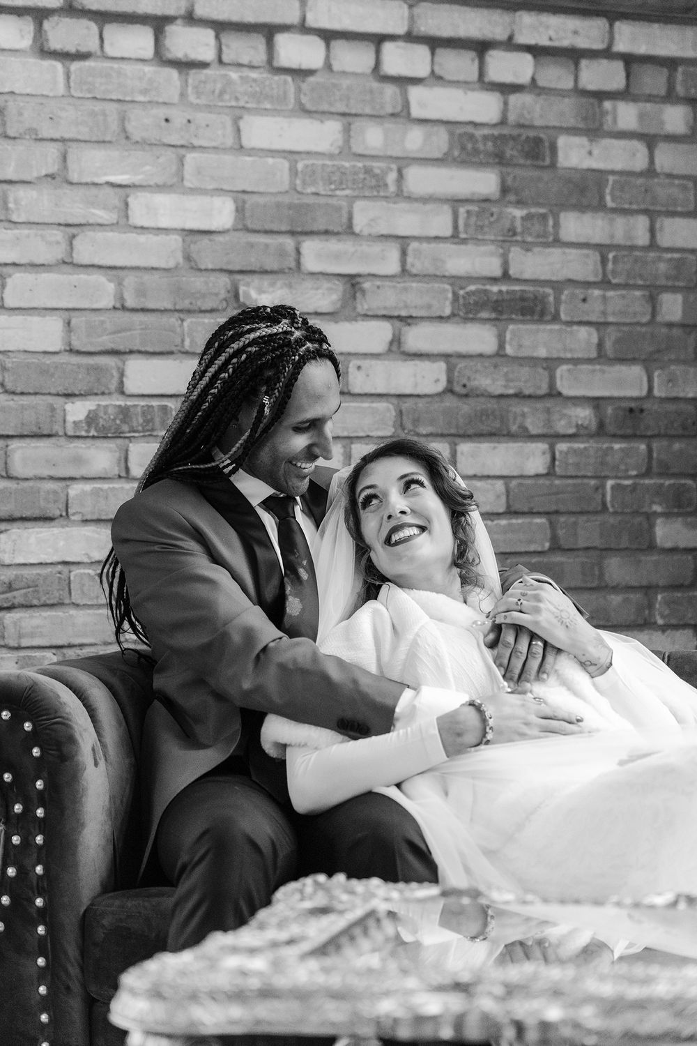 Black and white photo of interracial couple in wedding attire laughing together on a velvet sofa.