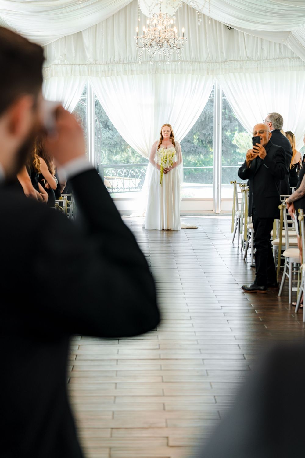 Bride walks down aisle in white gown holding bouquet in elegant draped wedding venue.