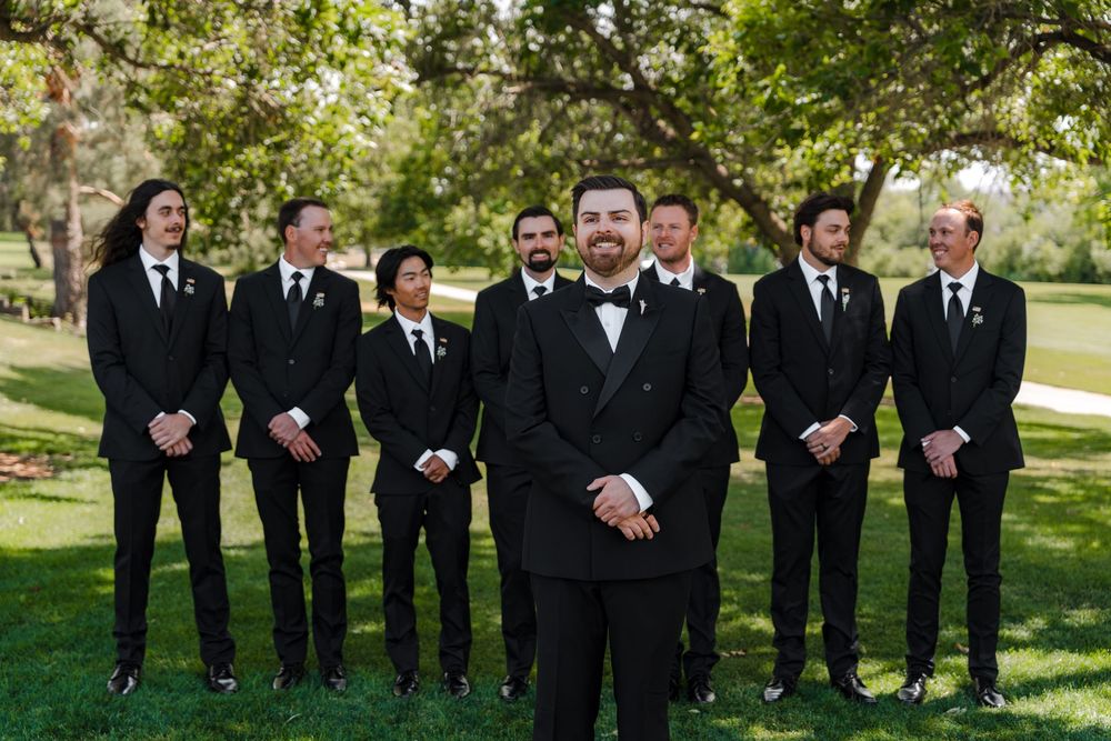 Groom in black tuxedo stands before groomsmen in matching black suits on a green lawn.