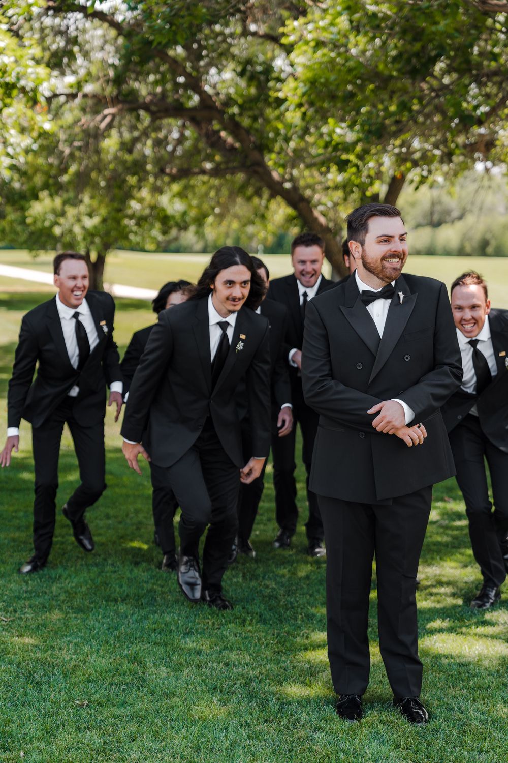 Groom in black tuxedo smiling as groomsmen in black suits walk behind him on green lawn.