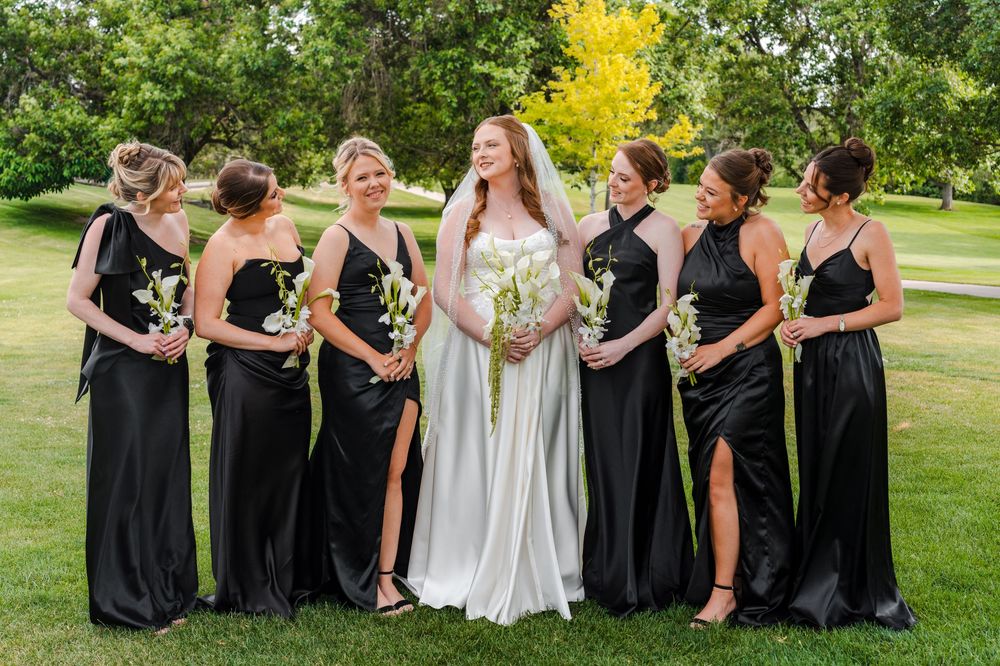 Bride in white gown poses with six bridesmaids in black dresses holding greenery bouquets on lush garden lawn.