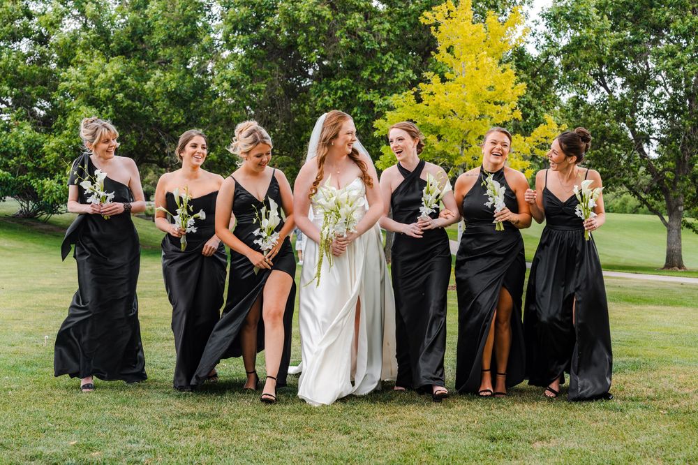 Bride in white gown walks with six bridesmaids in black dresses across lush green lawn.