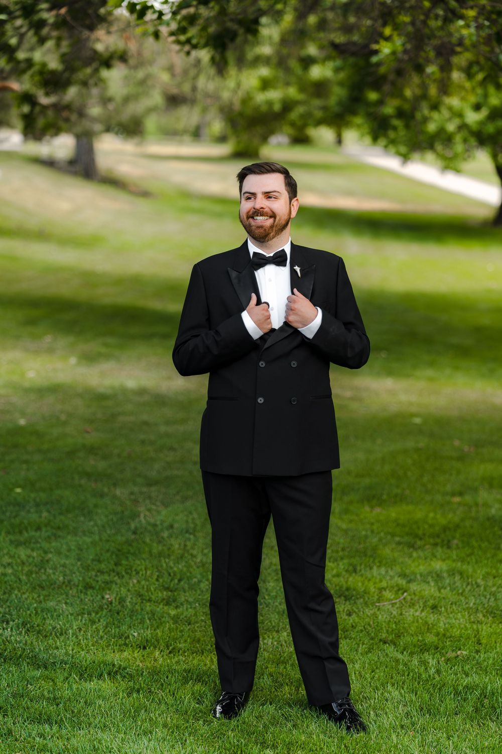 Smiling groom in black tuxedo and bow tie standing on green grass in outdoor park setting.