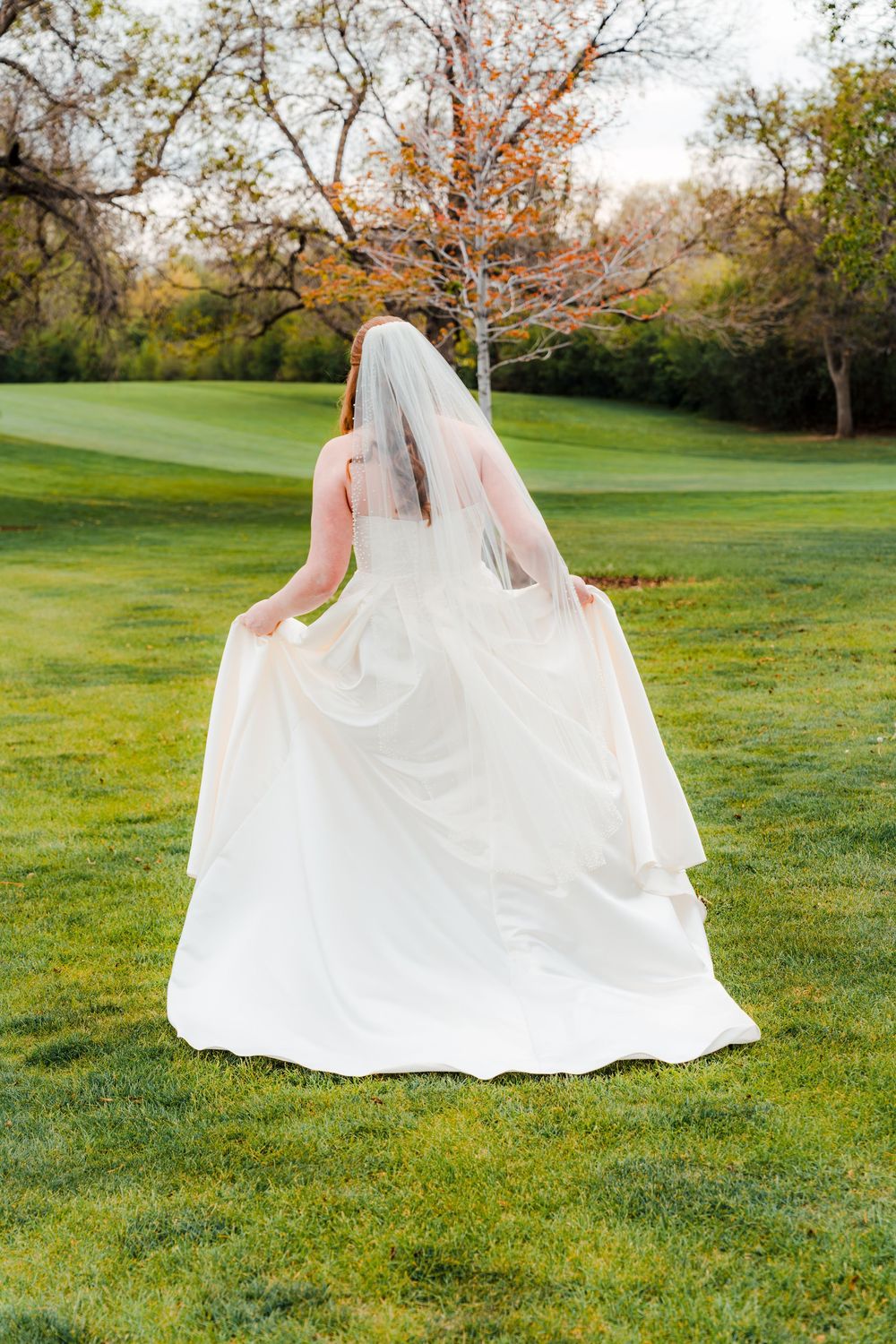 A bride in a white wedding gown and veil walks across a lush green lawn surrounded by autumn trees.