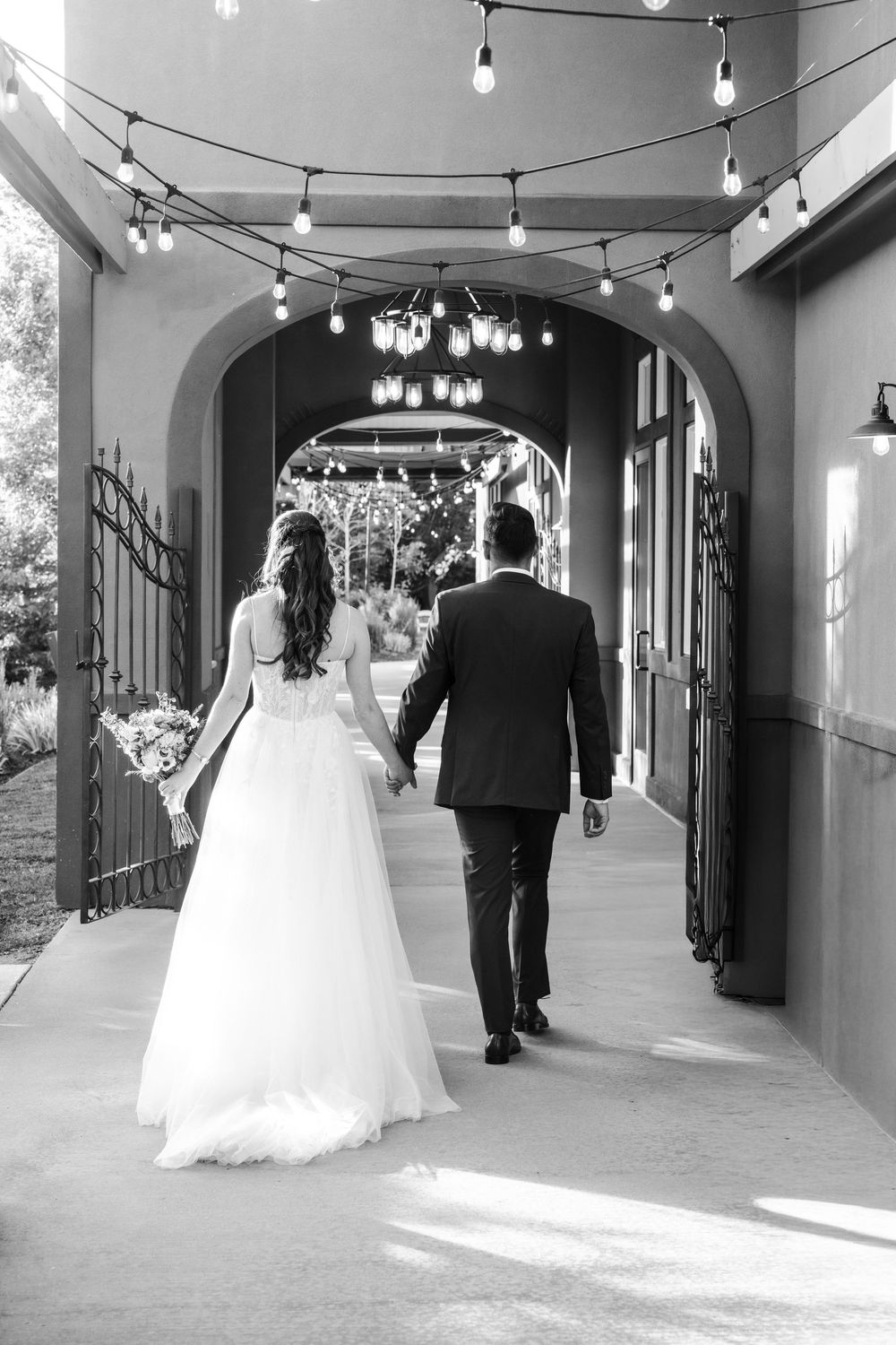 Black and white photo of bride and groom walking hand in hand through an elegant archway with string lights.