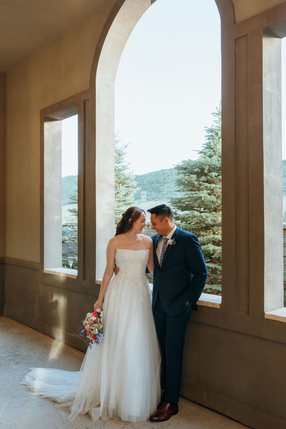 Bride and groom share an intimate moment under an arched window with mountain views at their elegant wedding venue.