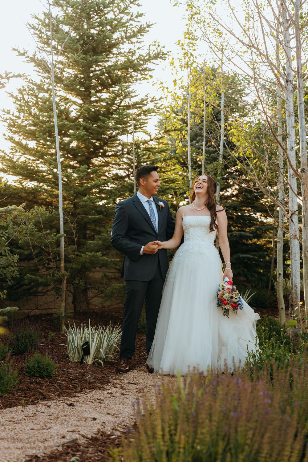 Happy bride and groom walk through a lush garden path, she laughs joyfully in a white tulle dress holding a vibrant bouquet.