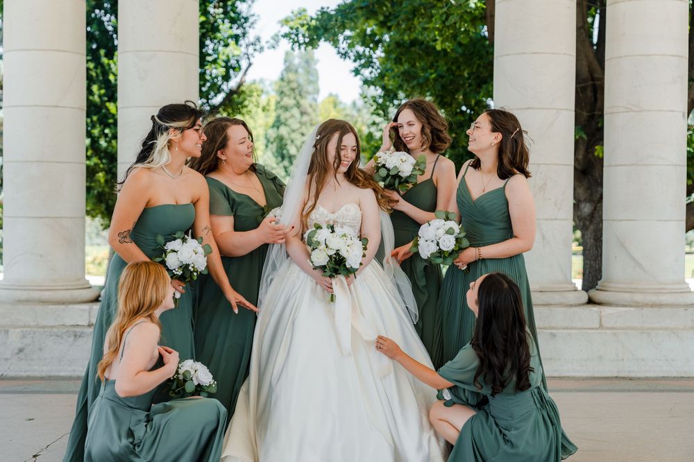 Bride in a white gown holding a bouquet, surrounded by six bridesmaids in green dresses with white flowers between stone columns.