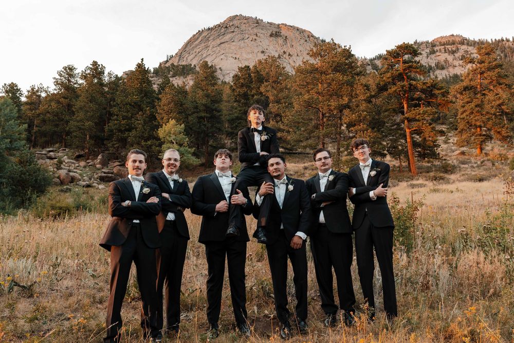 Groomsmen in black tuxedos pose with one lifted on shoulders in a mountain meadow setting.