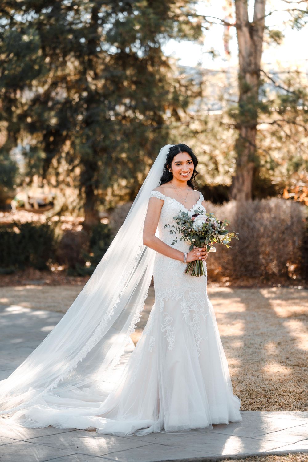 Bride in white lace mermaid gown with cathedral veil holds bouquet outdoors.