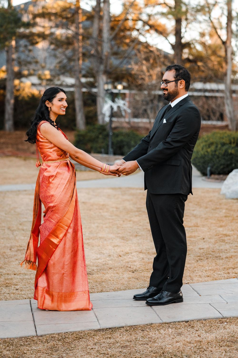 Couple holds hands outdoors, bride in orange saree and groom in black suit.