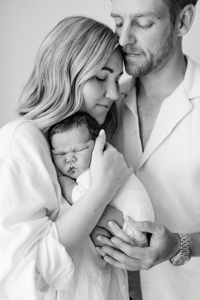 Intimate black and white photo showing new parents cuddled together holding their newborn baby against mums chest.