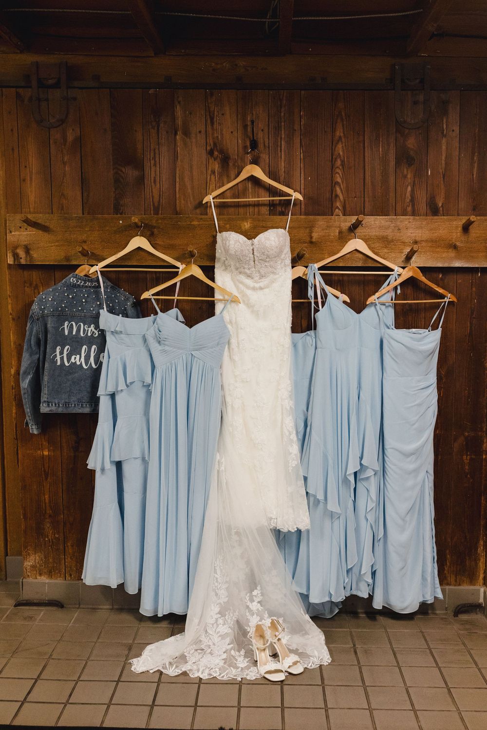 Wedding dress and light blue bridesmaid dresses hanging on wooden wall with bridal shoes displayed below.
