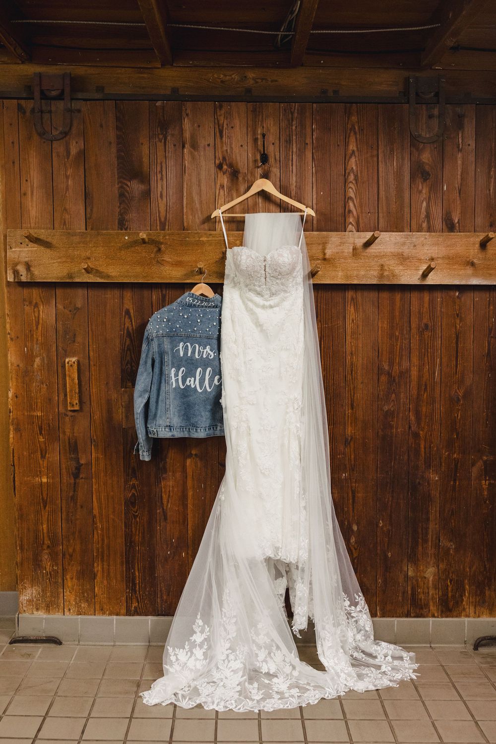 A white lace wedding dress and denim jacket hanging on wooden wall with rustic barn wood background.