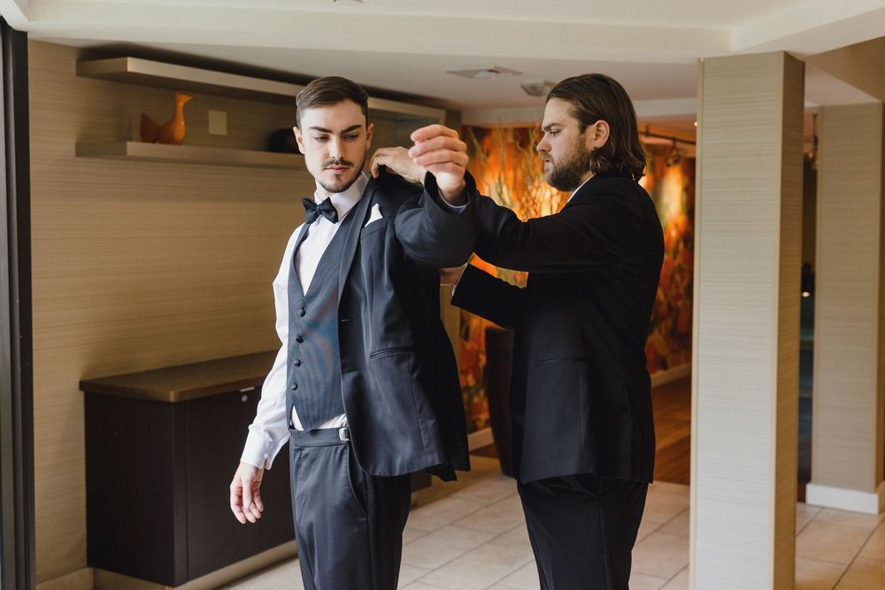A groom gets help adjusting his formal attire while getting ready in an elegant indoor setting.