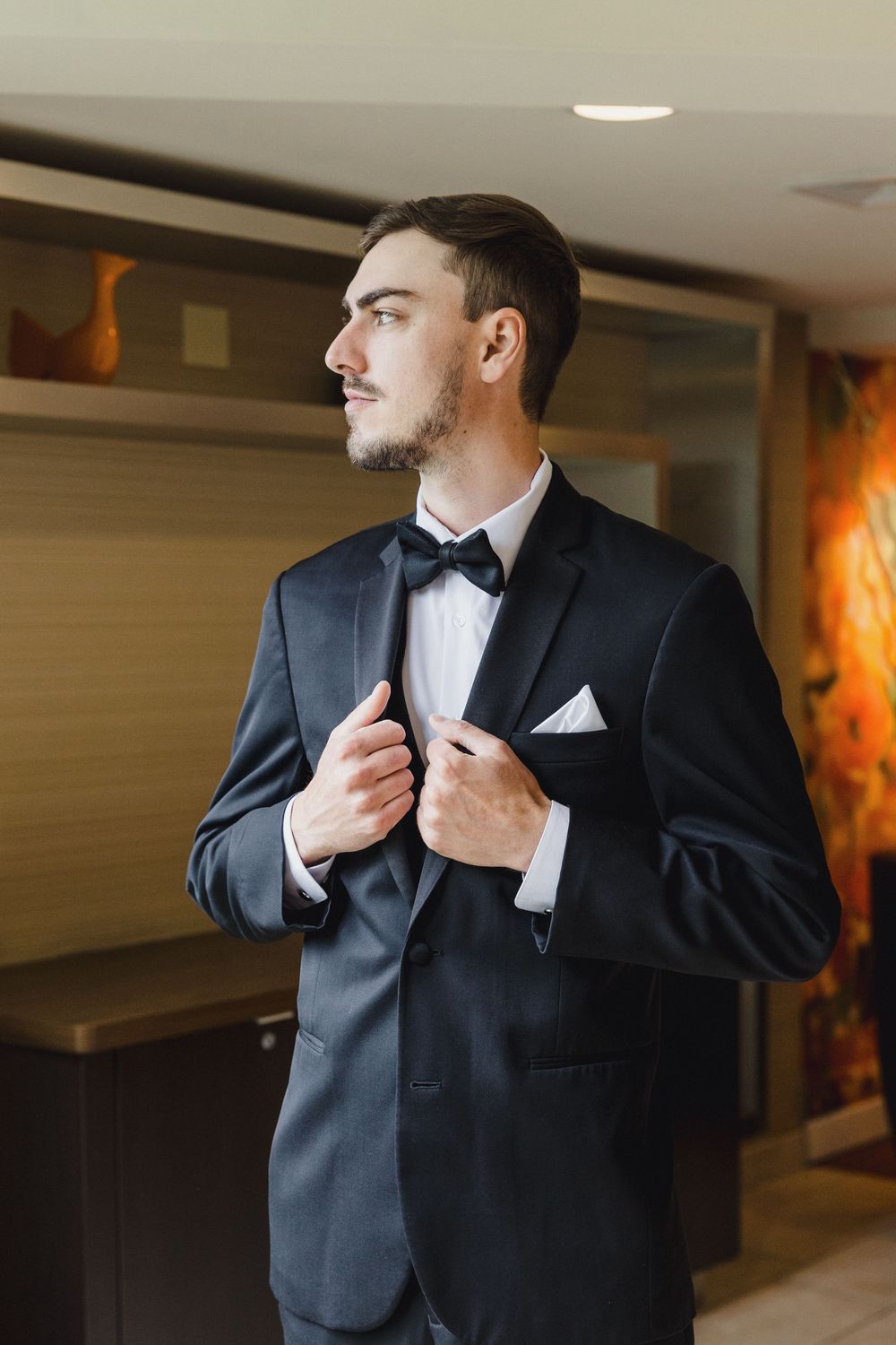 A groom in a classic black tuxedo and bow tie adjusts his jacket in a formal setting.