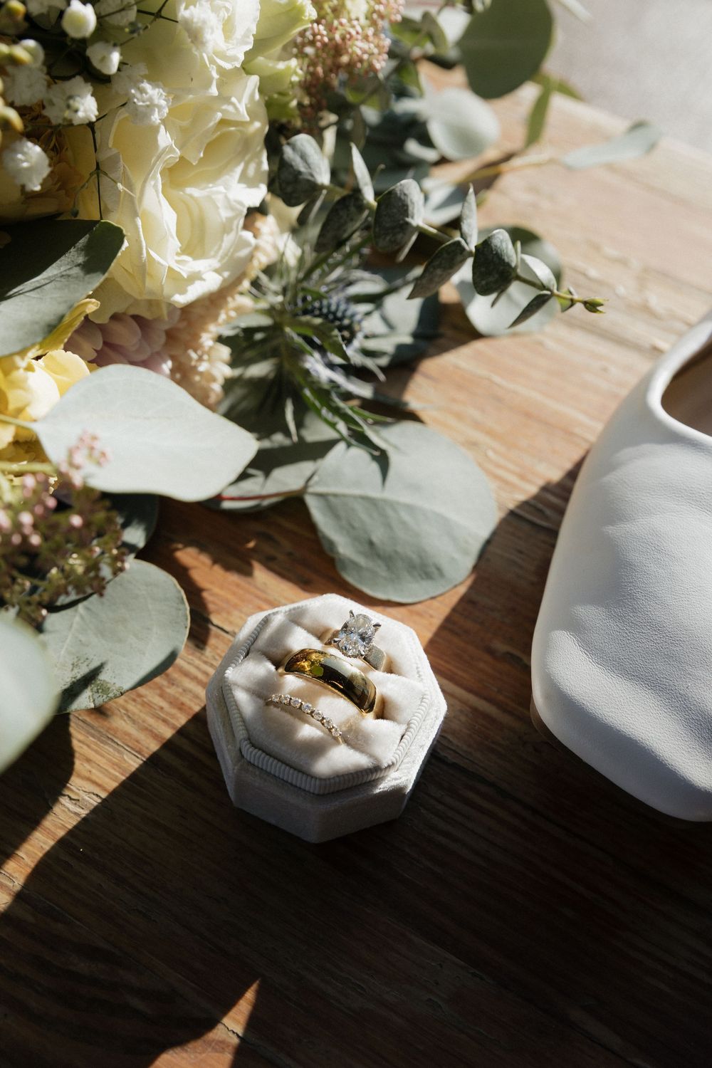 Wedding rings rest on white fabric cushions alongside delicate white floral arrangements and greenery on a dark wooden surface.