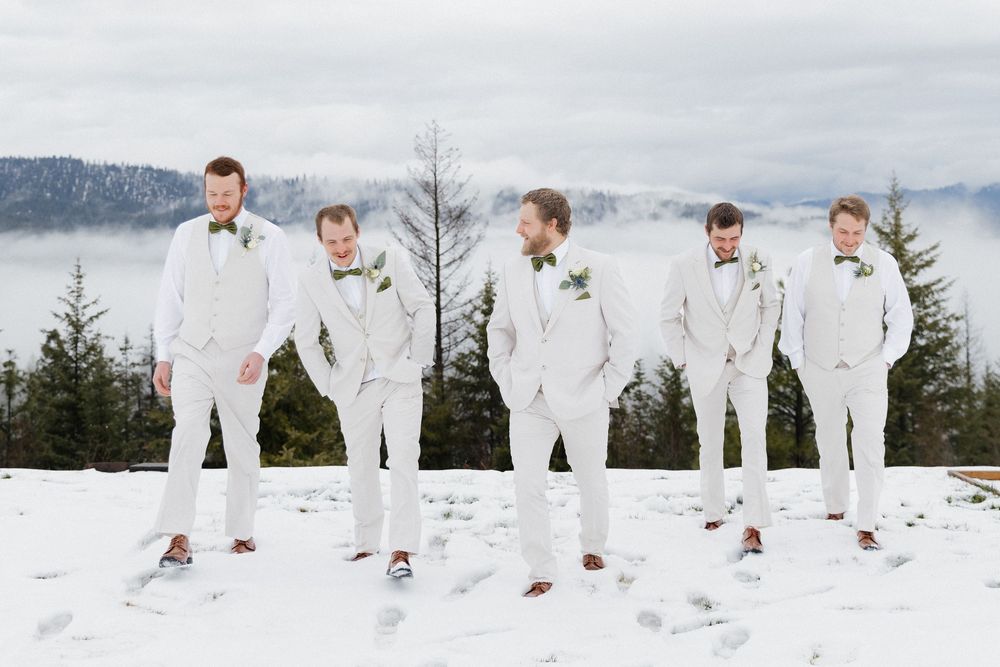 A group of five people in white suits stand in a snowy mountain landscape with evergreen trees in the background.