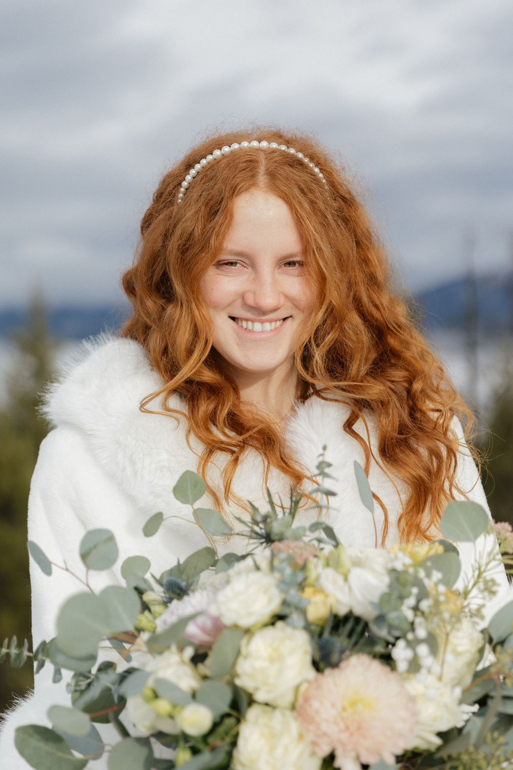 A bride in a white fur-trimmed dress holds a romantic bouquet of white and blush flowers with eucalyptus.