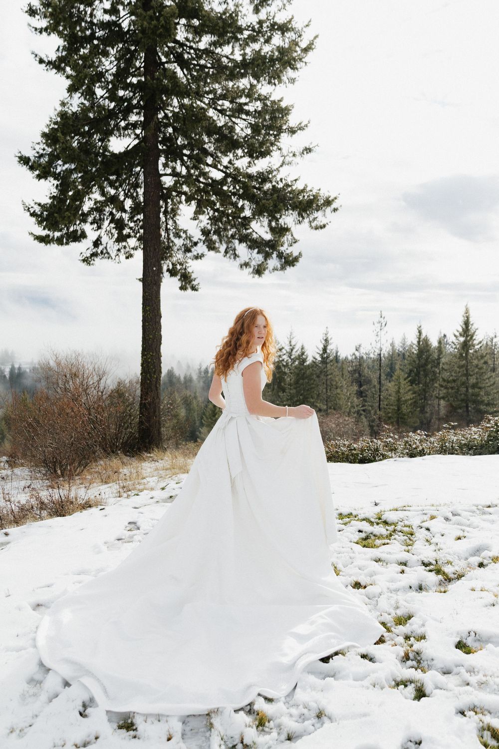 A white wedding dress flows elegantly in a snowy winter landscape with evergreen trees in the background.