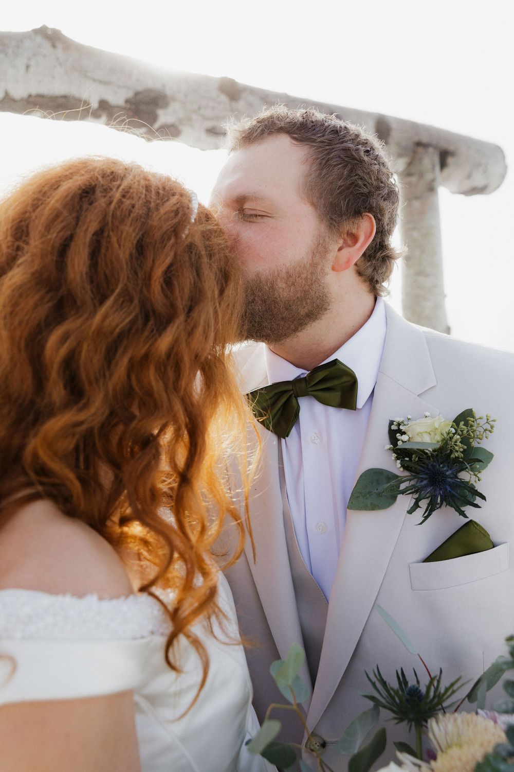 A romantic wedding couple shares an intimate kiss against a bright background.