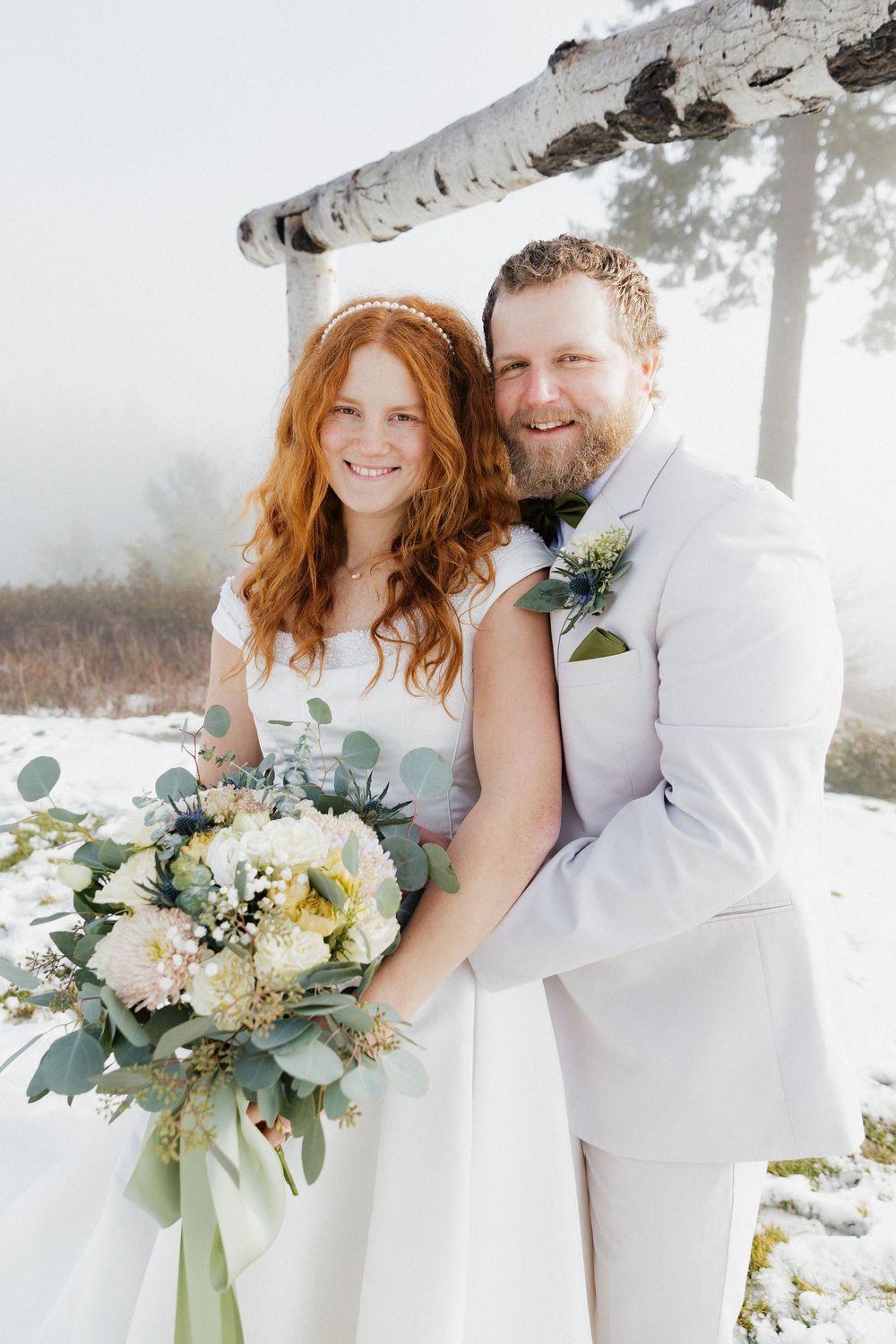 A couple poses together in winter wedding attire with a beautiful bouquet of flowers and eucalyptus against a snowy backdrop.