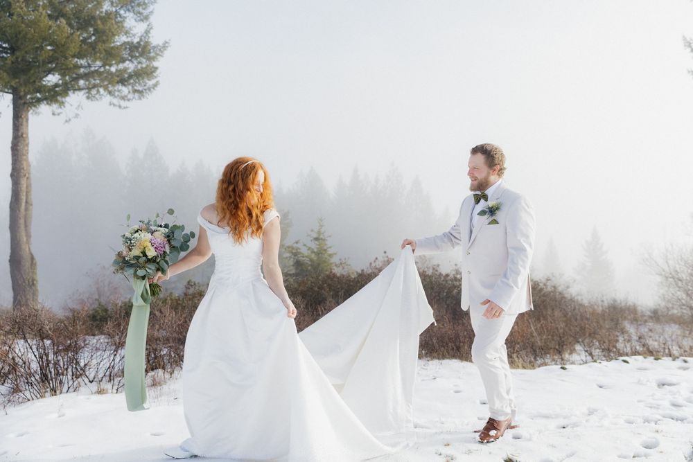 A winter wedding photo shoot in the snow with a white dress and suit against a misty forest backdrop.