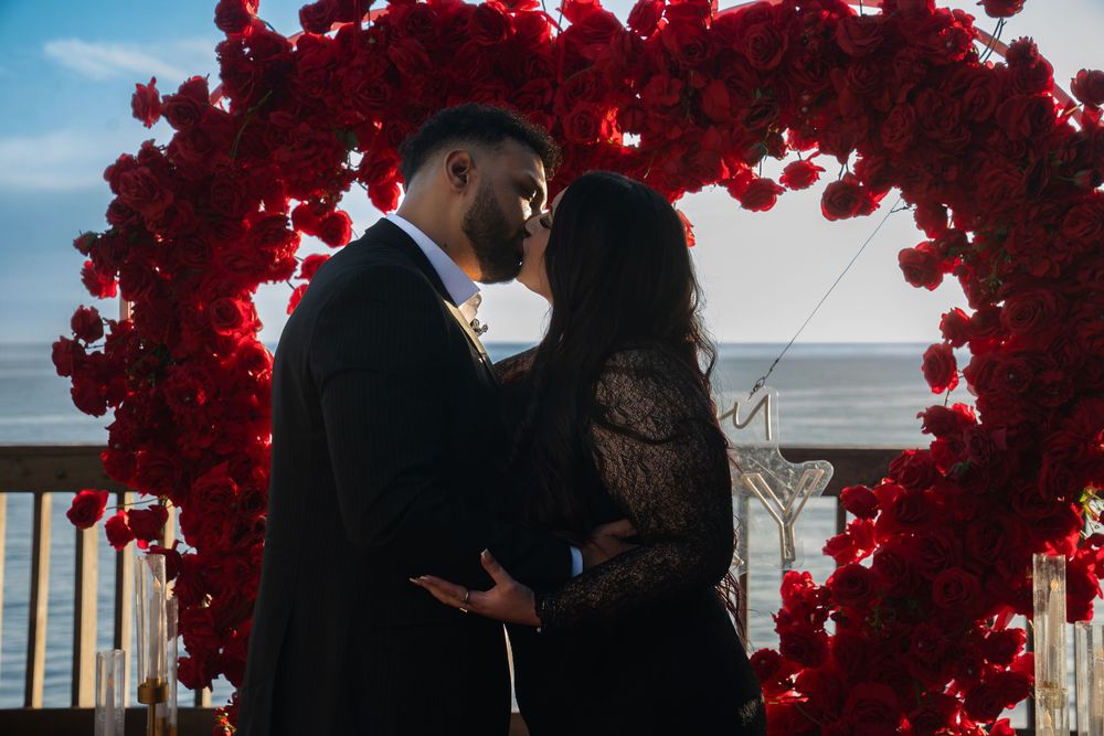 Red floral arch decorates an outdoor wedding ceremony venue with guests seated beneath a bright blue sky.