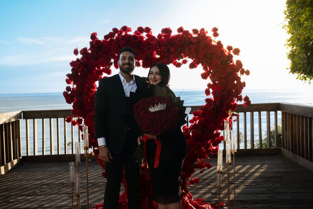 A romantic wedding ceremony under a red floral arch overlooking the ocean at sunset.