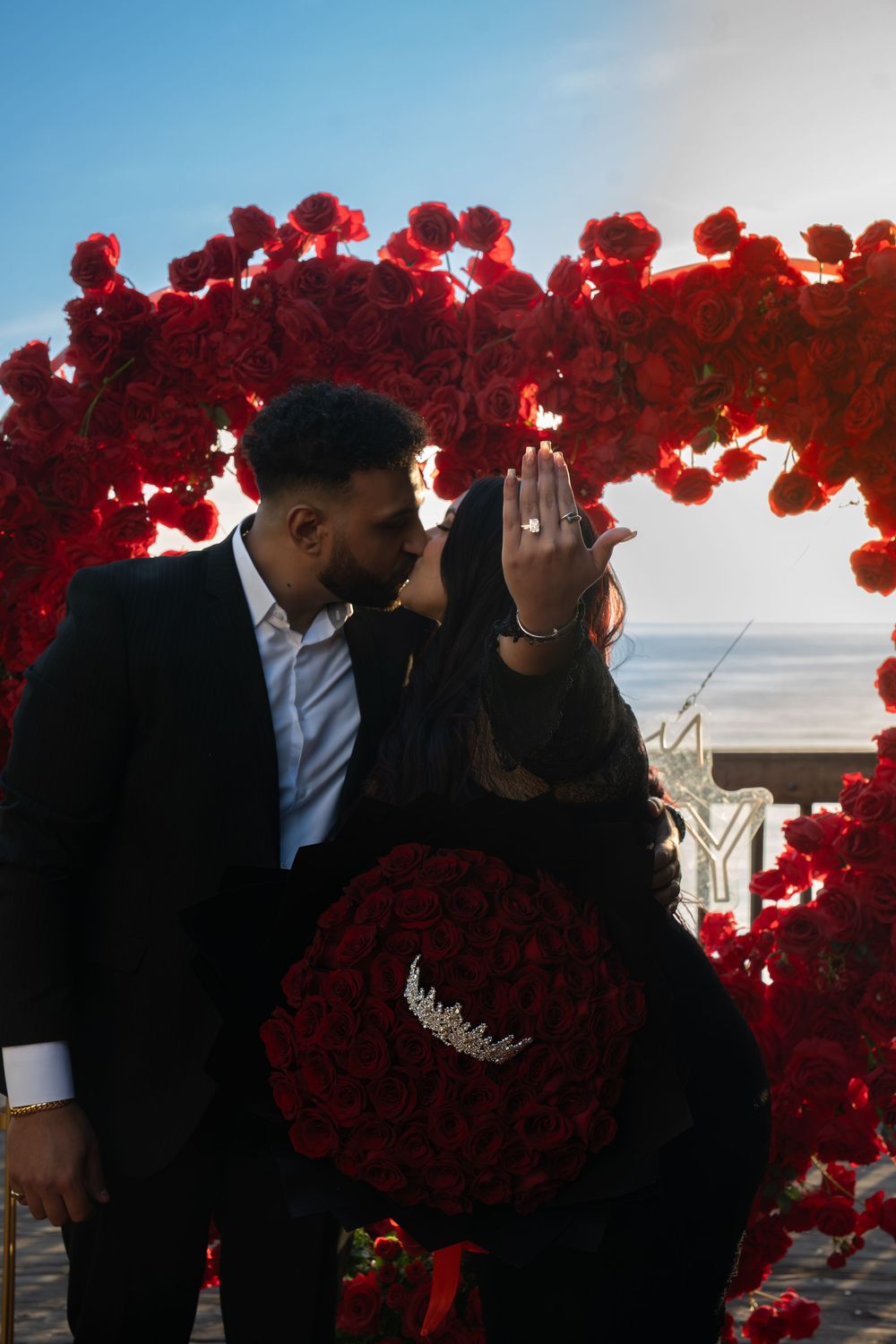 A couple shares a romantic moment against a sunset backdrop framed by vibrant red roses.