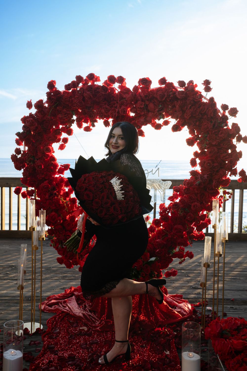 A romantic scene with red heart-shaped balloon arches on a wooden deck overlooking the ocean at sunset.