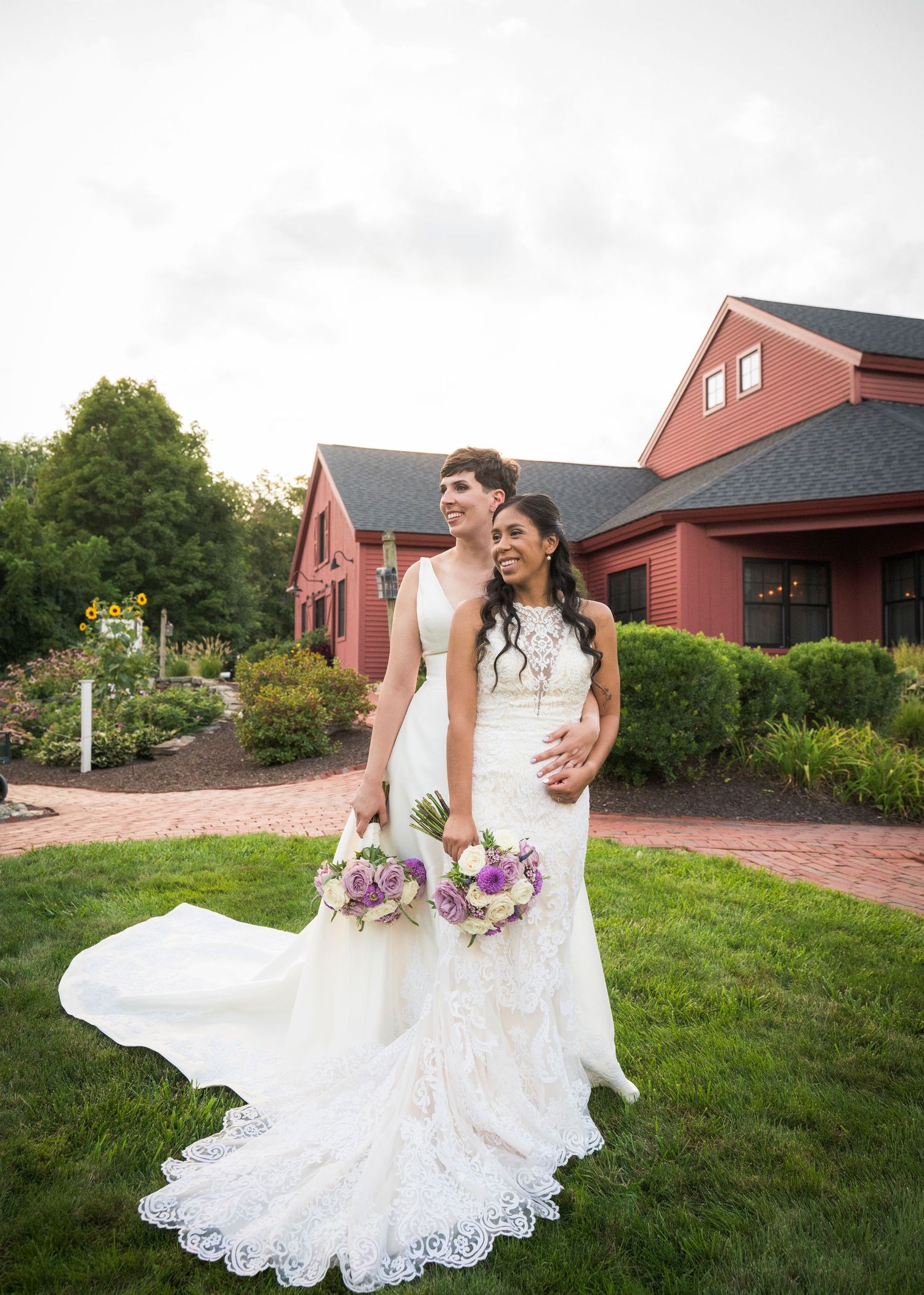 A wedding couple poses in front of a red barn venue with beautiful landscaping and purple floral bouquets.