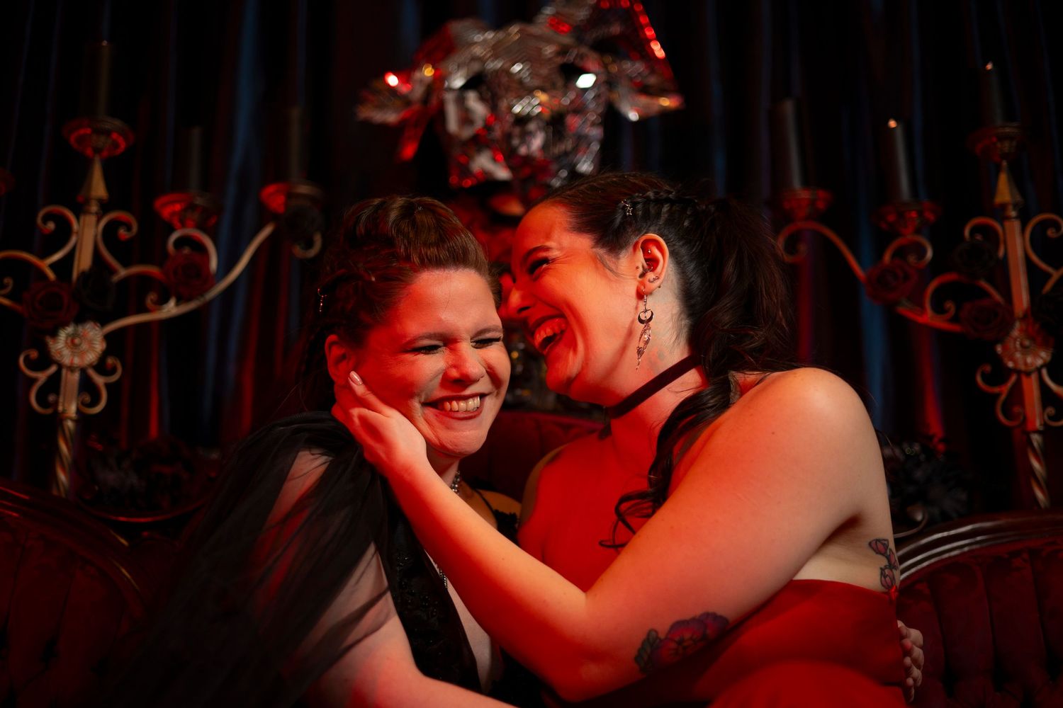 Two people share a joyful moment together in red lighting with ornate decorative elements in the background.