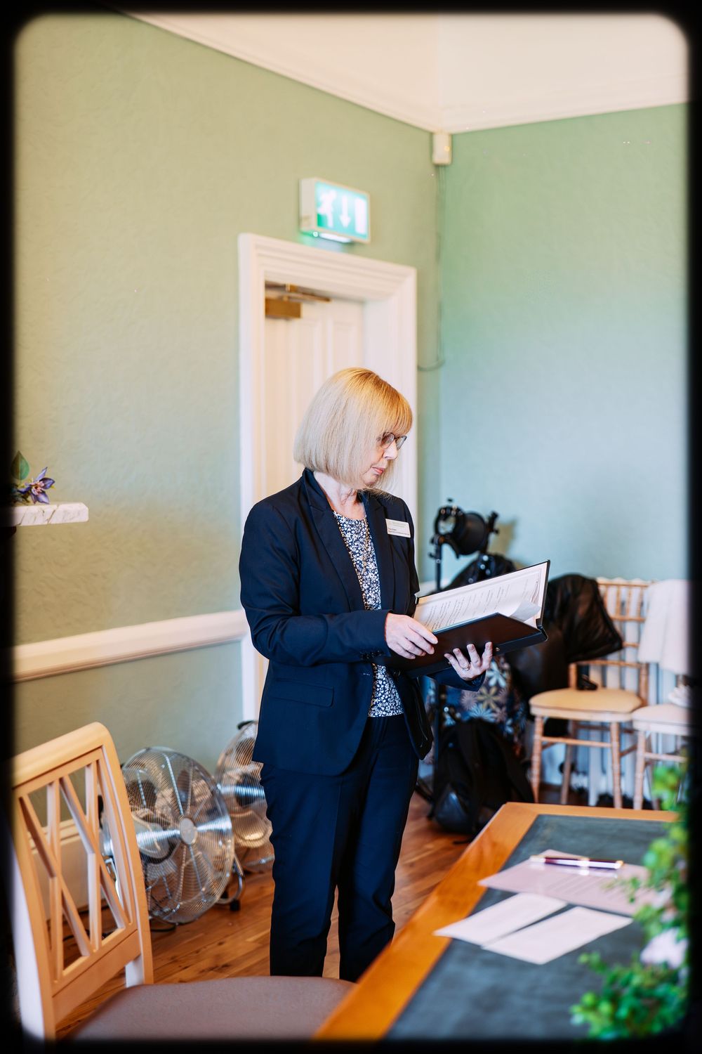 A person in a dark blazer reviews documents while standing at a wooden table in an office setting.