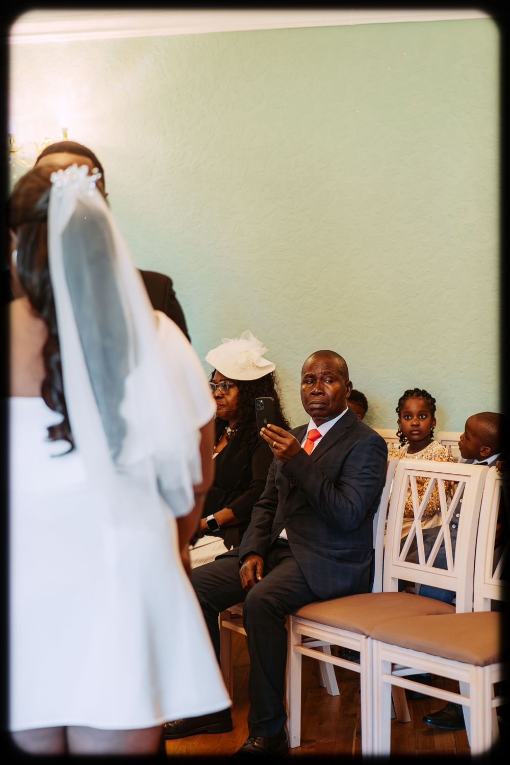 Wedding guests sit in white wooden chairs during a ceremony as a bride in a white dress and veil stands nearby.