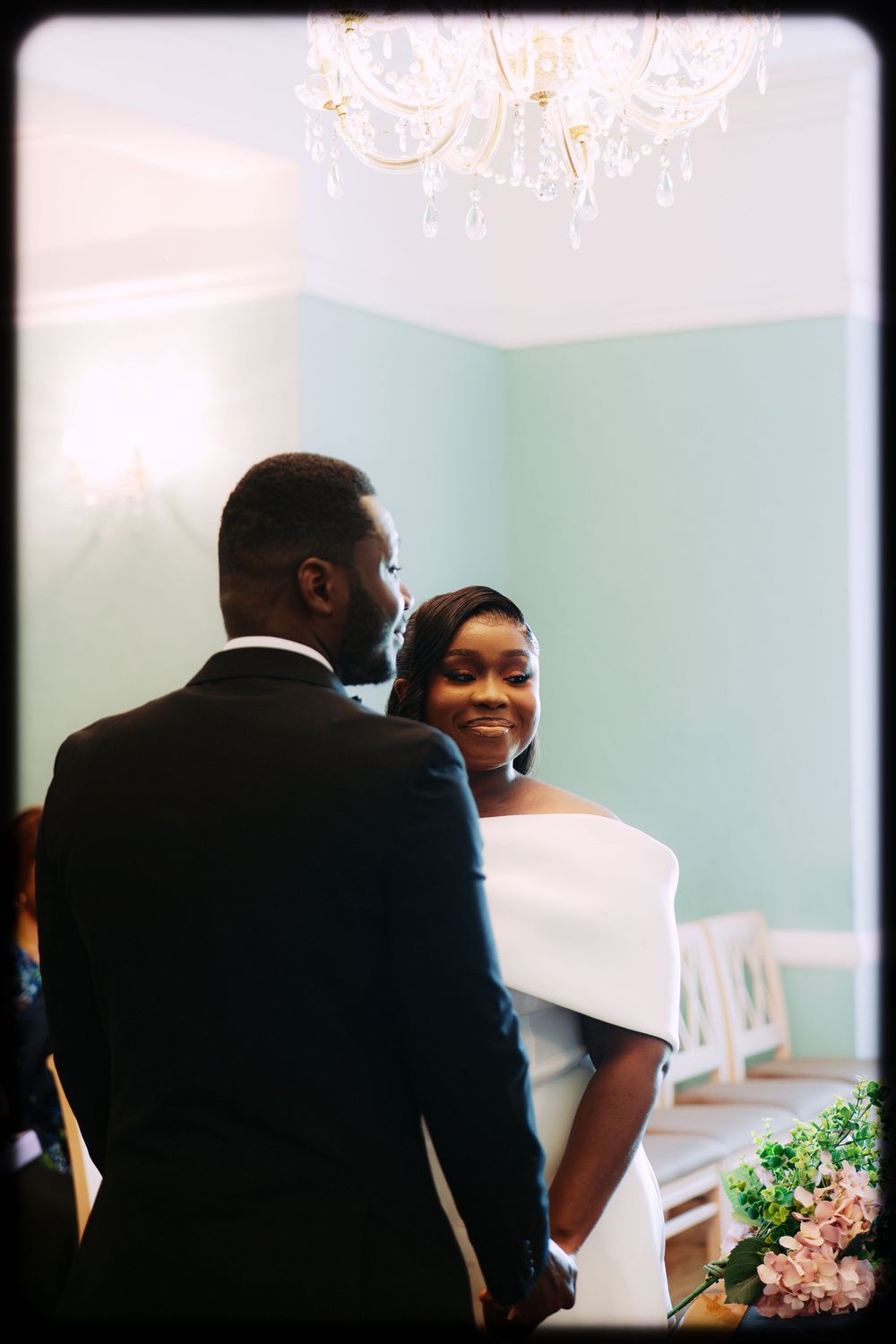 Wedding guests in formal attire gathered in an elegant room with mint green walls and a chandelier.